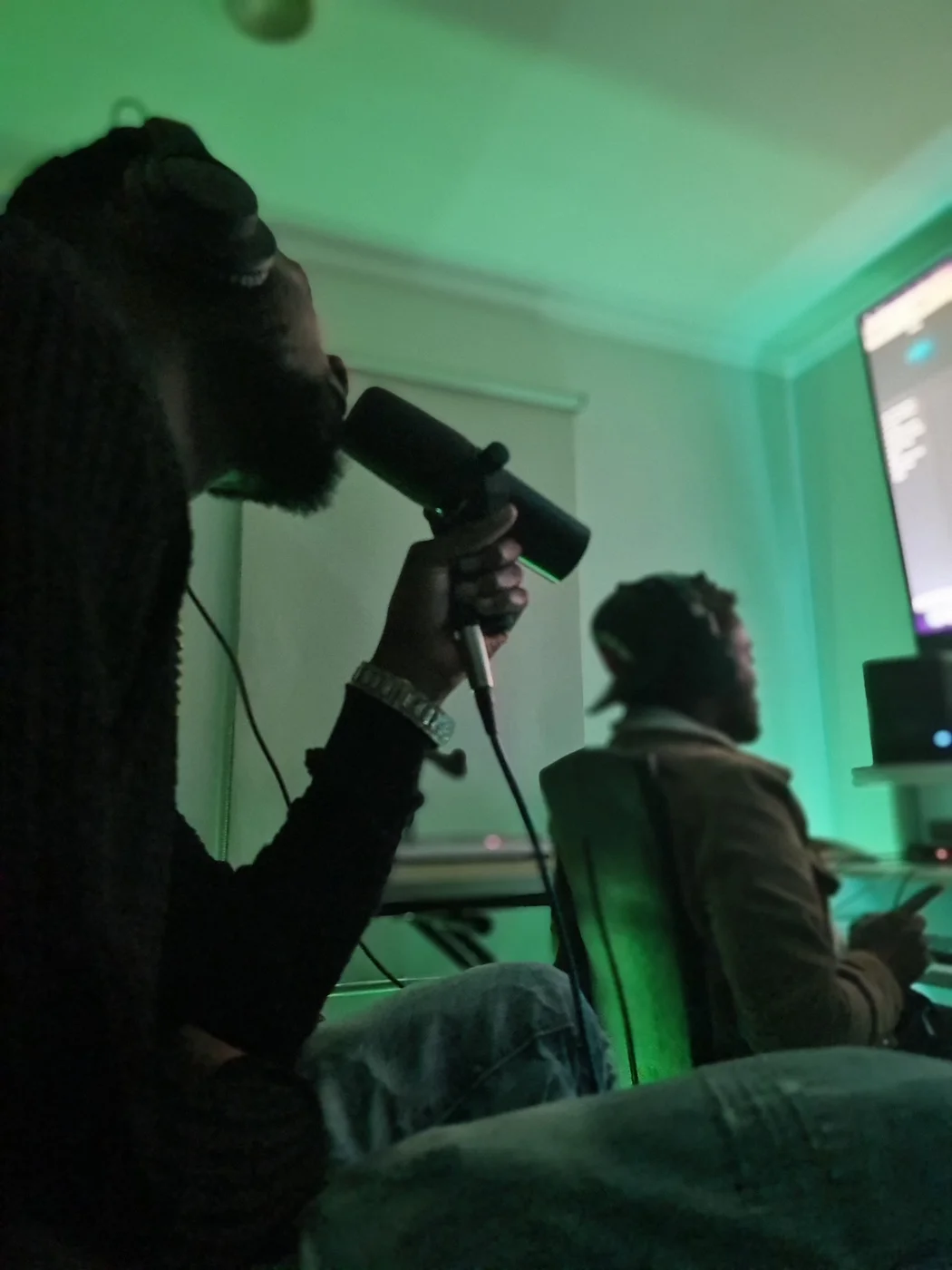 A vocalist recording with a microphone alongside a producer in a green-lit studio